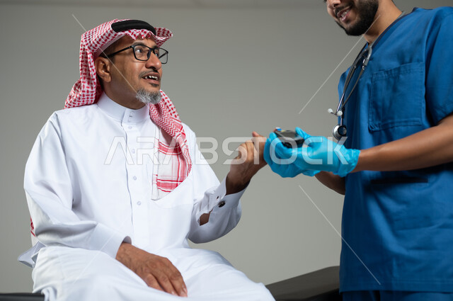 تقديم خدمات الفحص الطبي، العمل في القطاع الصحي في السعودية، بورتريه مقرب لممرض يرتدي قفازات الوقاية يقوم بفحص نسبة سكر الدم لرجل مريض عربي خليجي سعودي يرتدي الثوب التقليدي يجلس على سرير المرضى، خلفية رمادي، محتوى حساس، استخدام حساس
