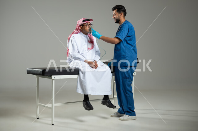 تقديم الخدمات الصحية  للمريض، المحافظة على ممر هوائي منتظم باستخدام جهاز التنفس الصناعي، معالجة امراض الربوة المزمنة، بورتريه لممرض عربي خليجي سعودي يرتدي اليونيفورم يقوم باعطاء ادوية الكحة لرجل مسن، الطول الكامل للجسم ، خلفية رمادية, محتوى حساس، اسخدام حساس