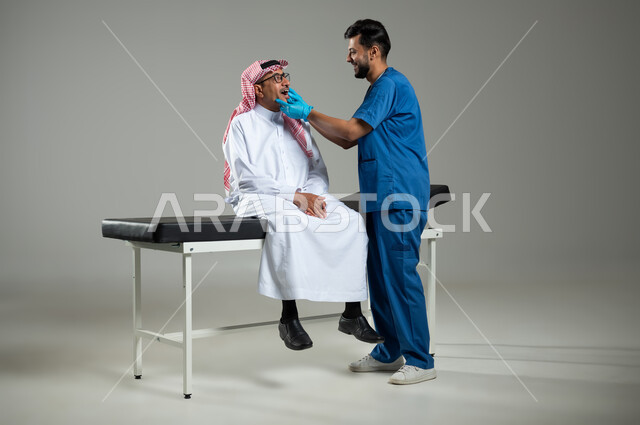تقديم الخدمات والرعاية الصحية، بورتريه مقرب لممرض عربي سعودي خليجي مبتسم يرتدي اليونيفورم والقفازات الطبية يقوم بفحص أسنان مريض عربي خليجي سعودي يرتدي الثوب التقليدي والشماغ، صورة الجسم بالطول الكامل، خلفية رمادية، محتوى حساس، استخدام حساس