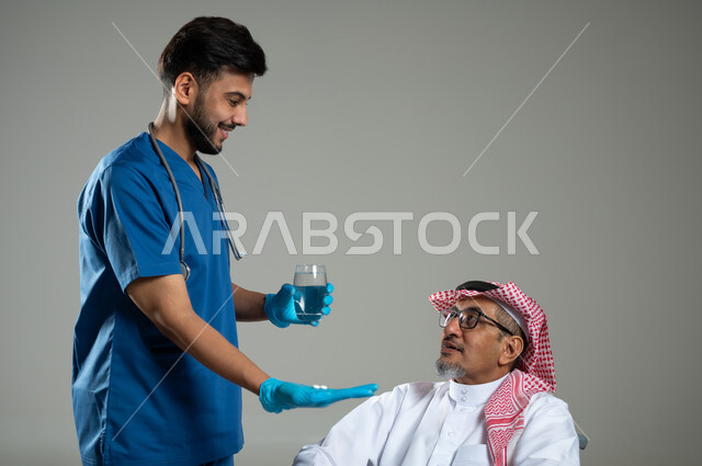 استخدام الدواء بوصفة طبية والاعتناء بالمريض، مفهوم الطب و الرعاية الصحية وتلقي العلاج، بورتريه مقرب لطبيب عربي سعودي خليجي مبتسم يرتدي اليونيفورم يعطي بعض الأدوية لمريض كبير بالسن يرتدي الشماغ والثوب التقليدي، خلفية رمادية، محتوى حساس، استخدام حساس .