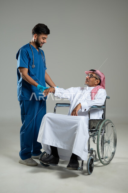 تقديم الخدمات الصحية، الاهتمام والاعتناء بمرضى ذوي الهمم، مفهوم الطب والرعاية الصحية، بورتريه مقرب لممرض يرتدي اليونيفورم يقدم العلاج الفيزيائي لرجل عربي خليجي سعودي مقعد يرتدي الثوب التقليدي، خلفية رمادية ، محتوى حساس، استخدام حساس