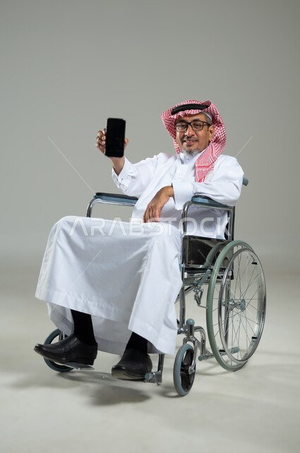 استخدام التقنيات الحديثة والمتطورة، النظر الى الكاميرا بتعابير السرور ,عرض شاشة سوداء فارغة عبر الجوال، بورتريه لرجل خليجي عربي سعودي يرتدي الثوب التقليدي والشماغ يجلس على كرسي متحرك يحمل بيده الهاتف المحمول، خلفية رمادية، استخدام حساس, محتوى حساس
