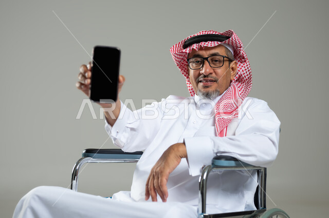النظر الى الكاميرا بتعابير السرور ,عرض شاشة سوداء فارغة عبر الجوال، بورتريه مقرب لرجل خليجي عربي سعودي يرتدي الثوب التقليدي والشماغ يجلس على كرسي متحرك يحمل بيده الهاتف المحمول، استخدام التقنيات الحديثة، خلفية رمادية، استخدام حساس, محتوى حساس