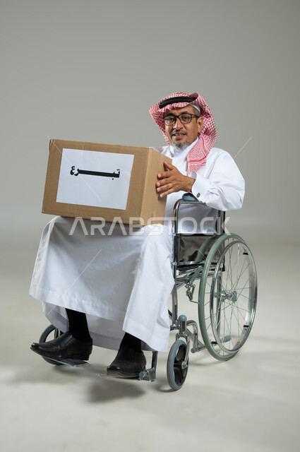 التبرع للجمعيات السعودية، التصدق بالفائض من المستلزمات، النظر الى الكاميرا بتعابير السرور، بورتريه لرجل مسن خليجي عربي سعودي يرتدي الثوب التقليدي يجلس على كرسي متحرك يحمل بيده صندوق التبرعات، خلفية رمادية، محتوى حساس، استخدام حساس