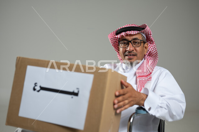 Not making the wheelchair an obstacle to doing good deeds, the concept of worship and getting closer to God, diligence in obedience and performing alms, looking at the camera with expressions of pleasure, a close-up portrait of an elderly Gulf Arab Saudi man wearing a formal uniform holding a donation box in his hand, Full length body image, gray background, sensitive content, sensitive use.