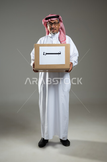 Donating and spending for the sake of God to Saudi associations, giving in charity surplus supplies, looking at the camera with expressions of pleasure, portrait of an elderly Gulf Arab Saudi man wearing formal dress holding a donation box in his hand, full length body, gray background, sensitive content, sensitive use