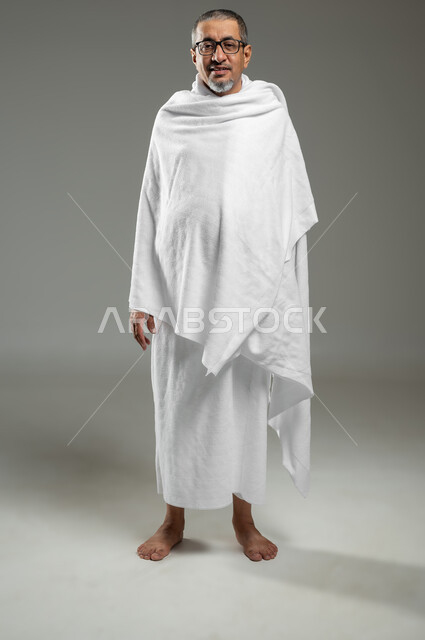 الاستعداد لأداء مناسك الحج، الوقوف باستقامة والنظر الى الكاميرا، بورتريه لرجل عربي خليجي سعودي مبتسم يرتدي لباس الاحرام يقوم بزيارة بيت الله الحرام في مكة المكرمة، الجسم بالطول الكامل، خلفية رمادية، الجسم بالطول الكامل، خلفية رمادية، محتوى حساس، استخدام حساس