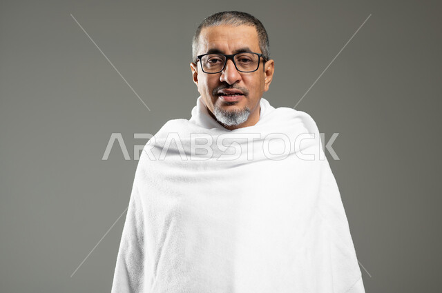 النظر إلى الكاميرا بإيماءات السرور، أداء مناسك الحج والعمره، بورتريه مقرب لحاج عربي سعودي خليجي مبتسم يرتدي ملابس الإحرام يقوم بزيارة بيت الله الحرام في مكة المكرمة، خلفية رمادية،  محتوى حساس، استخدام حساس