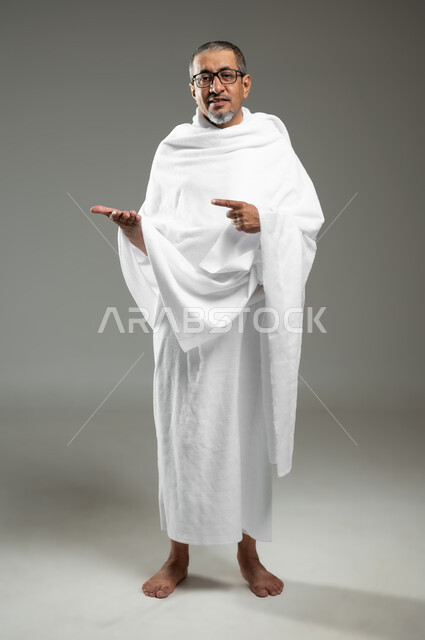 الإشارة بإصبع السبابة إلى شيء معين، أداء مناسك الحج والعمره، النظر إلى الكاميرا بإيماءات السرور ،بورتريه لرجل مسن عربي خليجي سعودي مبتسم يرتدي ملابس الإحرام يرفع يده للأعلى لعرض شيء ما، الجسم بالطول الكامل، خلفية رمادية، محتوى حساس، استخدام حساس