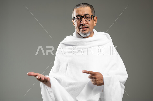 أداء مناسك الحج والعمره، النظر إلى الكاميرا بإيماءات السرور، الإشارة بإصبع السبابة إلى شيء معين، بورتريه لرجل مسن عربي خليجي سعودي مبتسم يرتدي ملابس الإحرام يرفع يده للأعلى لعرض شيء ما، خلفية رمادية،  محتوى حساس، استخدام حساس