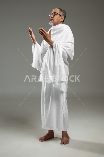 التقرب الى الله بالصلاة والدعاء والعبادة، أدعية وأذكار اسلامية , بورتريه لحاج مسلم عربي سعودي خليجي يرتدي ملابس الاحرام يدعو بخشوع وتضرع، أداء مناسك الحج والعمرة، الجسم بالطول الكامل، خلفية رمادية، محتوى حساس، استخدام حساس