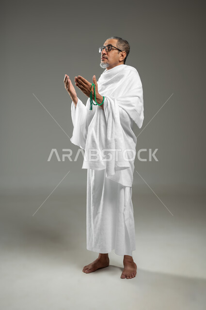 قراءة أذكار ما بعد الصلاة، التسبيح والتهليل وذكر الله تعالى، أداء مناسك الحج والعمرة، بورتريه لرجل مسلم عربي سعودي خليجي يرتدي ملابس الاحرام ويحمل بيده سبحة خضراء،قراءة الأذكار والأدعية، الجسم بالطول الكامل، خلفية رمادية، محتوى حساس، استخدام حساس