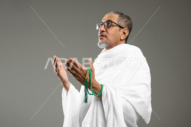 التسبيح والتهليل وذكر الله تعالى، أداء مناسك الحج والعمرة، بورتريه مقرب لرجل مسلم عربي سعودي خليجي يرتدي ملابس الاحرام ويحمل بيده سبحة خضراء،قراءة الأذكار والأدعية، خلفية رمادية، محتوى حساس، استخدام حساس