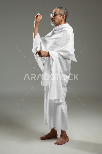رمي جمرة العقبة في يوم النحر، أداء مناسك الحج والعمرة، بورتريه لرجل مسن مسلم عربي سعودي خليجي يرتدي ملابس الاحرام يحمل بيده حصوى للقيام برجم ابليس، العبادة والتقرب الى الله، الجسم بالطول الكامل، خلفية رمادية، محتوى حساس، استخدام حساس