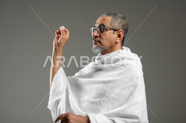 Performing the rituals of Hajj and Umrah, throwing Jamarat al-Aqaba on the Day of Sacrifice, a close-up portrait of an elderly Saudi Arab Gulf Muslim man wearing Ihram clothes, holding a pebble in his hand to stone Satan, worship and getting closer to God, gray background