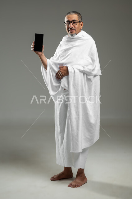 عرض شاشة سوداء فارغة عبر الجوال،  النظر الى الكاميرا بتعابير السرور, بورتريه لرجل مسلم خليجي عربي سعودي يرتدي ملابس الاحرام يحمل بيده الهاتف المحمول، أداء مناسك الحج والعمرة، استخدام التقنيات الحديثة، الجسم بالطول الكامل، خلفية رمادية، محتوى حساس، استخدام حساس