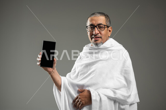 النظر الى الكاميرا بتعابير السرور ,عرض شاشة سوداء فارغة عبر الجوال، بورتريه مقرب لرجل مسلم خليجي عربي سعودي يرتدي ملابس الاحرام يحمل بيده الهاتف المحمول، أداء مناسك الحج والعمرة، استخدام التقنيات الحديثة، خلفية رمادية، استخدام حساس، محتوى حساس