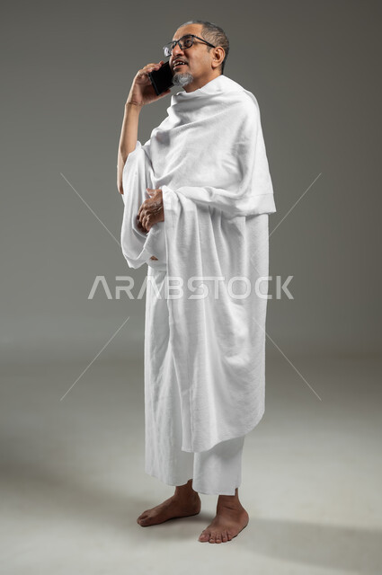 استخدام تقنيات حديثة، التواصل مع العائلة والأصدقاء، زيارة بيت الله الحرام في مكة المكرمة، بورتريه لحاج عربي خليجي سعودي مبتسم يرتدي ملابس الاحرام يقوم بإجراء مكالمة صوتية عبر الهاتف المحمول , الجسم بالطول الكامل، خلفية رمادية، محتوى حساس، استخدام حساس