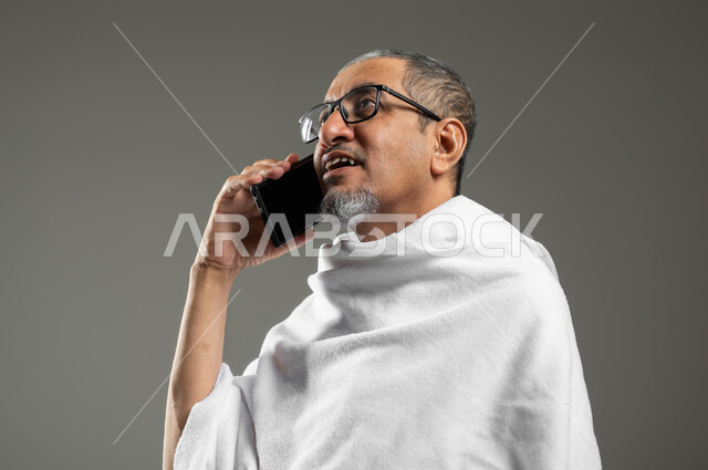 زيارة بيت الله الحرام في مكة المكرمة، استخدام تقنيات حديثة، التواصل مع العائلة والأصدقاء، بورتريه مقرب لحاج عربي خليجي سعودي مبتسم يرتدي ملابس الاحرام يقوم بإجراء مكالمة صوتية عبر الجوال , أداء مناسك الحج والعمرة ، خلفية رمادية، محتوى حساس، استخدام حساس