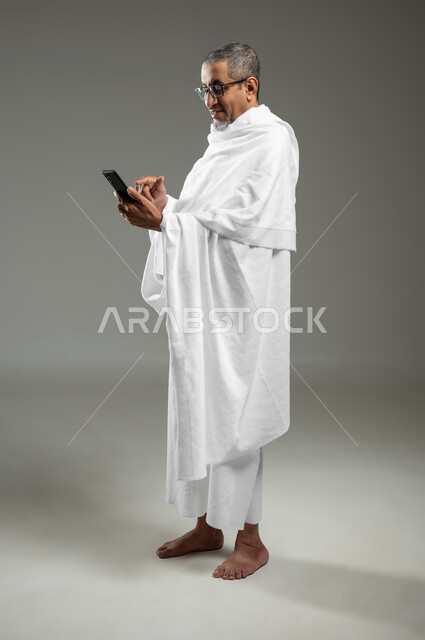 استخدام الهاتف المحمول، تطبيق مرشد الحج الالكتروني، أداء مناسك الحج والعمرة، بورتريه لحاج مسلم خليجي عربي سعودي مبتسم يرتدي ملابس الإحرام يستخدم الجوال، الجسم بالطول الكامل، خلفية رمادية، محتوى حساس، استخدام حساس
