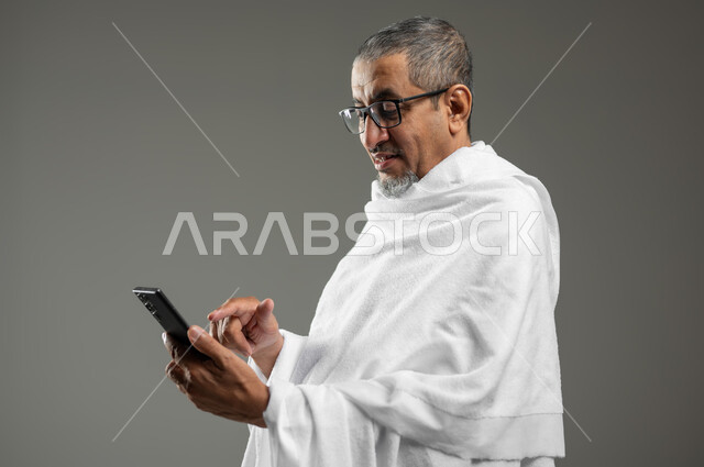 تطبيق الادعية والاذكار، استخدام تطبيقات الارشادات لأداء مناسك الحج والعمرة، بورتريه مقرب لحاج مسلم خليجي عربي سعودي مبتسم يرتدي ملابس الإحرام يستخدم الهاتف المحمول، خلفية رمادية، محتوى حساس، استخدام حساس