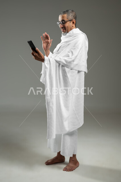 القاء التحية من خلال شاشة الهاتف المحمول، اجراء مكالمة فيديو باستخدام الجوال،  بورتريه لحاج مسلم خليجي عربي سعودي مبتسم يرتدي ملابس الاحرام يقوم بالتقاط صورة سيلفي تذكارية من خلال الهاتف المحمول، الجسم بالطول الكامل، خلفية رمادية، محتوى حساس، استخدام حساس