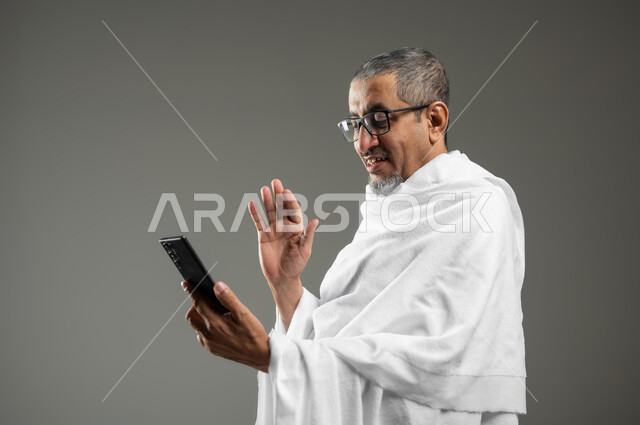 اجراء مكالمة فيديو باستخدام الهاتف المحمول، استخدام أجهزة تقنية حديثة، أداء مناسك الحج والعمرة، بورتريه مقرب لرجل عربي خليجي سعودي مبتسم يرتدي ملابس الاحرام يقوم بالتقاط صورة سيلفي تذكارية من خلال الهاتف المحمول، خلفية رمادية، محتوى حساس، استخدام حساس