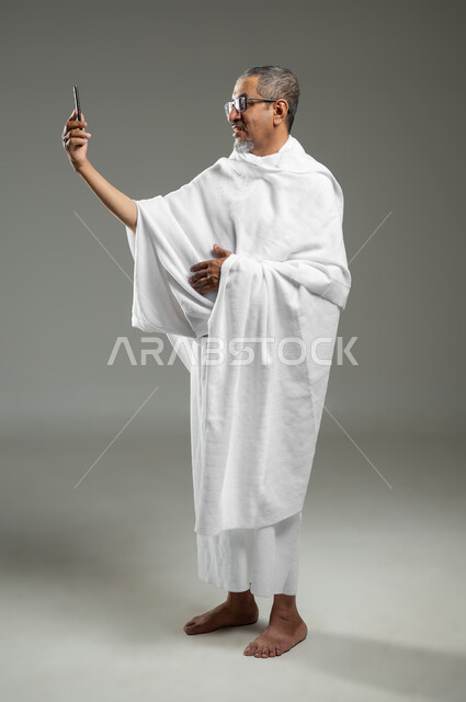 استخدام أجهزة تقنية حديثة، توثيق اللحظات السعيدة في مكة المكرمة،  أداء مناسك الحج والعمرة، بورتريه لرجل عربي خليجي سعودي يرتدي ملابس الاحرام ويقوم بالتقاط صور سيلفي تذكارية عبر الهاتف المحمول، الجسم بالطول الكامل، خلفية رمادية، محتوى حساس، استخدام حساس