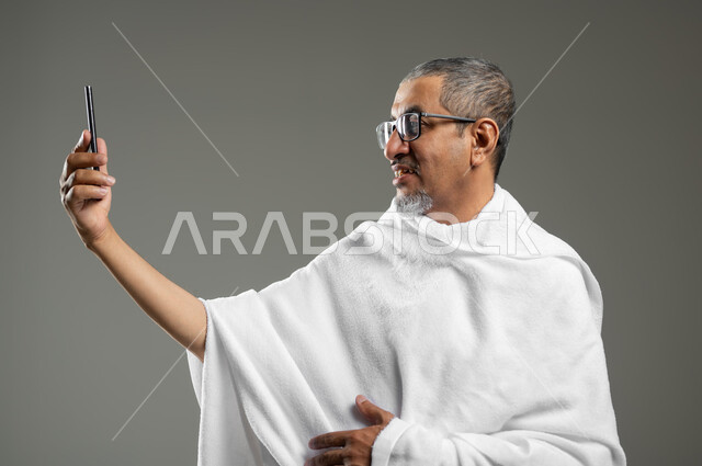 توثيق اللحظات السعيدة في مكة المكرمة، استخدام أجهزة تقنية حديثة، أداء مناسك الحج والعمرة، بورتريه مقرب لرجل عربي خليجي سعودي يرتدي ملابس الاحرام ويقوم بالتقاط صور سيلفي تذكارية عبر الهاتف المحمول، خلفية رمادية، محتوى حساس، استخدام حساس