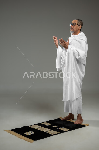 أداء مناسك الحج والعمرة، التقرب الى الله من خلال الاجتهاد في العبادة والحفاظ على الطاعات، بورتريه لرجل عربي سعودي خليجي يرتدي لباس الإحرام يرفع يديه ويدعو الله بإيماءات تدل على التوسل والتضرع، الجسم بالطول الكامل، خلفية رمادية، محتوى حساس، استخدام حساس