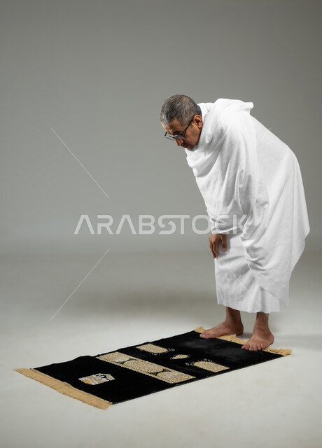 التقرب الى الله من خلال الاجتهاد في العبادة وأداء مناسك الحج والعمرة، بورتريه لحاج عربي خليجي سعودي يرتدي ملابس الإحرام يؤدي فريضة الصلاة ، خلفية رمادية، محتوى حساس، استخدام حساس