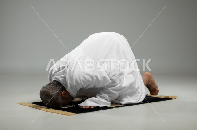 السجود على سجادة الصلاة, أداء الفرائض الدينية و مناسك الحج والعمرة، مفهوم العبادة والتقرب إلى الله تعالى ، بورتريه لحاج عربي خليجي سعودي يرتدي ملابس الإحرام يؤدي فريضة الصلاة ، خلفية رمادية، محتوى حساس، استخدام حساس