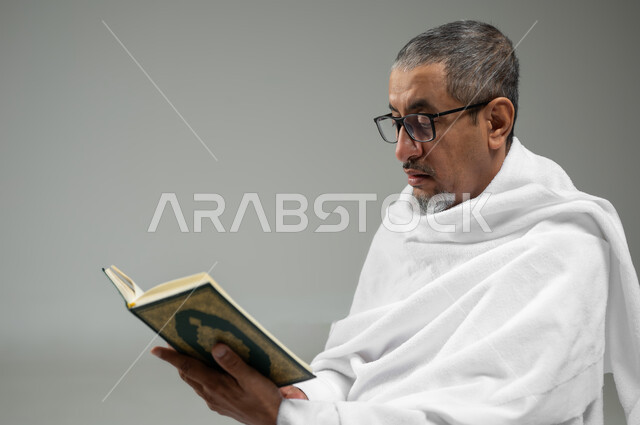 قراءة كتاب الله، مفهوم العبادة والتقرب الى الله, بورتريه مقرب لرجل عربي سعودي خليجي يرتدي لباس الاحرام يحمل بيده كتاب القرآن الكريم ، أداء مناسك الحج والعمرة، خلفية رمادية