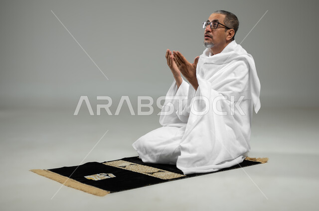الجلوس على سجادة الصلاة، العبادة والتقرب الى الله، أداء مناسك الحج والعمرة، بورتريه مقرب لحاج مسلم عربي سعودي خليجي يرتدي ملابس الاحرام يرفع يديه للأعلى بإيماءات الدعاء، أداء مناسك الحج والعمرة، خلفية رمادية، محتوى حساس، استخدام حساس
