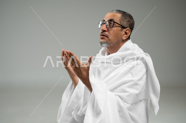 التقرب الى الله بالصلاة والعبادة، بورتريه مقرب لحاج مسلم عربي سعودي خليجي يرتدي ملابس الاحرام يرفع يديه للأعلى بإيماءات الدعاء، أداء مناسك الحج والعمرة، خلفية رمادية، محتوى حساس، استخدام حساس