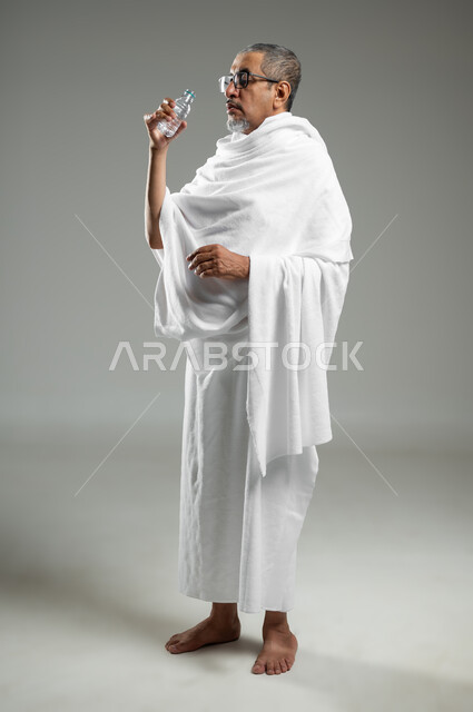 الارتواء بماء زمزم، الاكثار من السوائل للحفاظ على الجسم من الجفاف، أداء مناسك الحج والعمرة، بورتريه لرجل عربي سعودي خليجي يرتدي زي الاحرام يقوم بشرب المياه العذبة، الجسم بالطول الكامل، خلفية رمادية، محتوى حساس، استخدام حساس