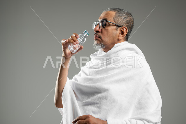 Performing Hajj and Umrah rituals, plenty of fluids to keep the body from dehydration, close-up portrait of a Saudi Gulf Arab man wearing the Ihram dress drinking fresh water, quenching himself with Zamzam water, gray background, sensitive content, sensitive use