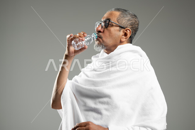 الاكثار من السوائل للحفاظ على الجسم من الجفاف، أداء مناسك الحج والعمرة، بورتريه مقرب لرجل عربي سعودي خليجي يرتدي زي الاحرام يقوم بشرب المياه  العذب، الارتواء بماء زمزم، خلفية رمادية، محتوى حساس، استخدام حساس