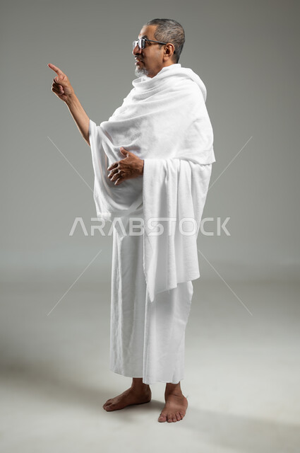 الوقوف باستقامة و النظر إلى مكان معين، بورتريه لرجل مسلم عربي سعودي خليجي يرتدي ملابس الاحرام يشير بأصبع السبابة نحو الأعلى الى مكان ما، أداء مناسك الحج والعمرة، الجسم بالطول الكامل، خلفية رمادية، محتوى حساس، استخدام حساس