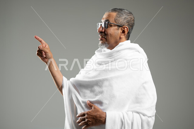 Standing straight and looking at a specific place, a close-up portrait of a Saudi Arabian Gulf Muslim man wearing Ihram clothing, pointing with his index finger upwards to a specific place, performing Hajj and Umrah rituals, gray background, sensitive content