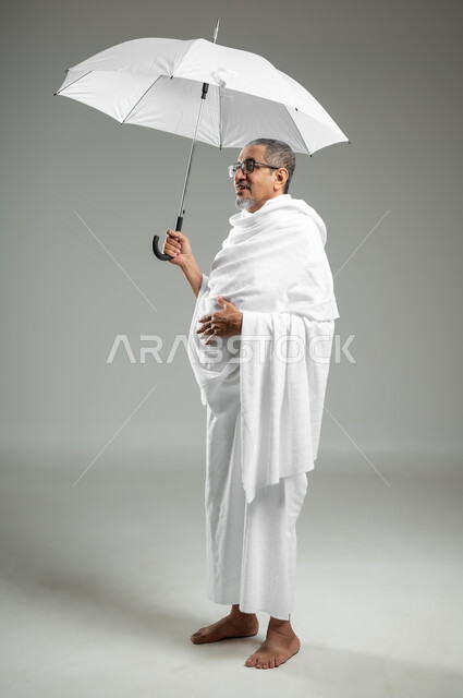 أداء مناسك الحج والعمرة، العبادة والتقرب إلى الله بالأعمال الصالحة، بورتريه لرجل عربي سعودي خليجي يرتدي ملابس الاحرام يحمل بيده مظلة للحماية من أشعة الشمس، الجسم بالطول الكامل، خلفية رمادية،محتوى حساس، استخدام حساس