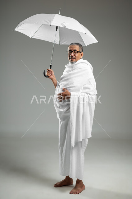 الذهاب لأداء مناسك الحج، العبادة والتقرب إلى الله بالأعمال الصالحة، بورتريه لرجل عربي سعودي خليجي يرتدي ملابس الاحرام يحمل بيده مظلة للحماية من أشعة الشمس، الجسم بالطول الكامل، خلفية رمادية، محتوى حساس، استخدام حساس