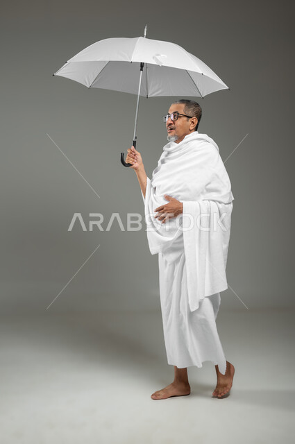 Walking and going to perform Hajj and Umrah rituals, worship and getting closer to God with good deeds, portrait of a Saudi Gulf Arab man wearing Ihram clothes holding a sun umbrella in his hand, full-length body image, gray background, sensitive content, sensitive use