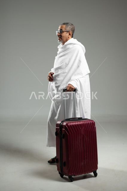 أداء فريضة الحج و العمرة، المشي والذهاب لزيارة بيت الله الحرام, بورتريه لرجل كبير السن عربي خليجي سعودي يرتدي ملابس الاحرام و يمسك بحقيبة سفر حمراء,  الجسم بالطول الكامل، خلفية رمادية، استخدام حساس, محتوى حساس