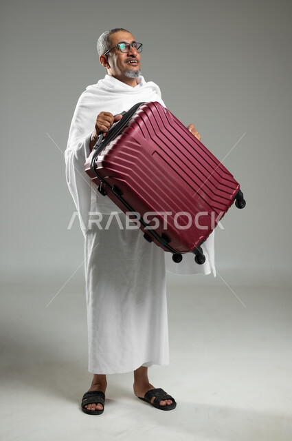 التوجه والانطلاق لزيارة بيت الله الحرام, بورتريه  لرجل كبير  السن عربي خليجي سعودي يرتدي ملابس الاحرام و يحمل حقيبة سفر حمراءبيديه , أداء فريضة الحج و العمرة, الجسم بالطول الكامل, خلفية رمادية، محتوى حساس، استخدام حساس