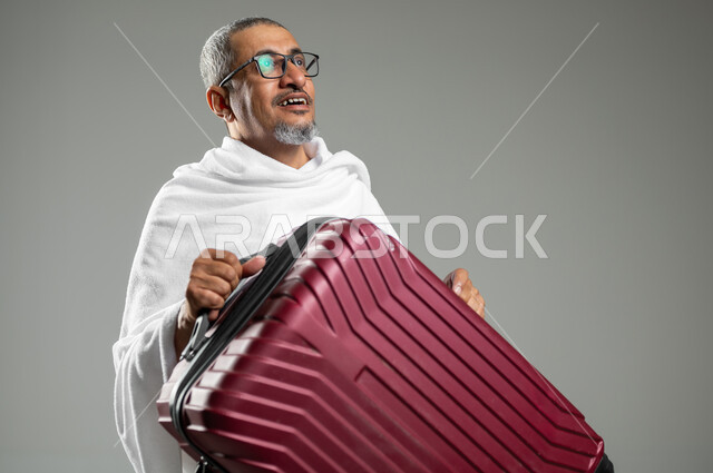 الاستعداد للتوجه و الانطلاق لزيارة بيت الله الحرام, بورتريه مقرب لرجل كبير  السن عربي خليجي سعودي يرتدي ملابس الاحرام و يحمل حقيبة سفر حمراء, الذهاب لآداء فريضة الحج و العمرة , ايماءات السعادة والحماس للوصول لمكة المكرمة, خلفية رمادية, محتوى حساس, استخدام حساس