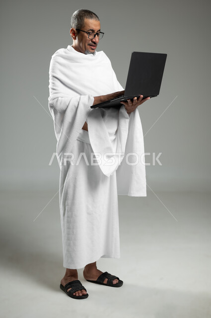 استخدام الأجهزة التقنية الحديثة في تصفح مواقع الارشادات لأداء مناسك الحج والعمرة، بورتريه لرجل كبير السن مسلم خليجي عربي سعودي مبتسم يرتدي ملابس الاحرام يحمل بيده الحاسوب المحمول، الجسم بالطول الكامل, خلفية رمادية,محتوى حساس, استخدام حساس