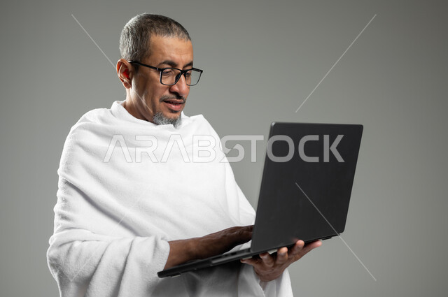 الأجهزة التقنية الحديثة, تصفح مواقع الارشادات لأداء مناسك الحج والعمرة، بورتريه مقرب لرجل كبير السن  مسلم خليجي عربي سعودي مبتسم يرتدي ملابس الاحرام يحمل بيده جهاز الحاسوب المحمول، خلفية رمادية, محتوى حساس , استخدام حساس
