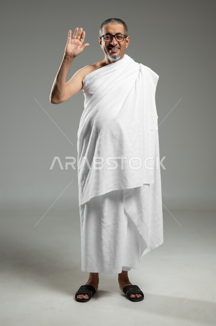 النظر إلى الكاميرا بإيماءات السرور، أداء مناسك الحج والعمرة، بورتريه لرجل عربي سعودي خليجي مبتسم يرتدي ملابس الاحرام ويرفع يده للأعلى لإلقاء التحية والسلام، الجسم بالطول الكامل، خلفية رمادية، استخدام حساس, محتوى حساس