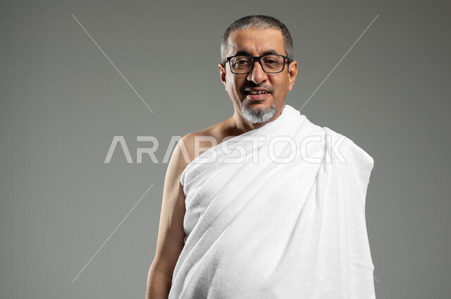 أداء مناسك الحج والعمرة، النظر إلى الكاميرا بإيماءات السرور، بورتريه مقرب لحاج عربي سعودي خليجي مبتسم يرتدي ملابس الإحرام يقوم بزيارة بيت الله الحرام ، خلفية رمادية،  استخدام حساس, محتوى حساس